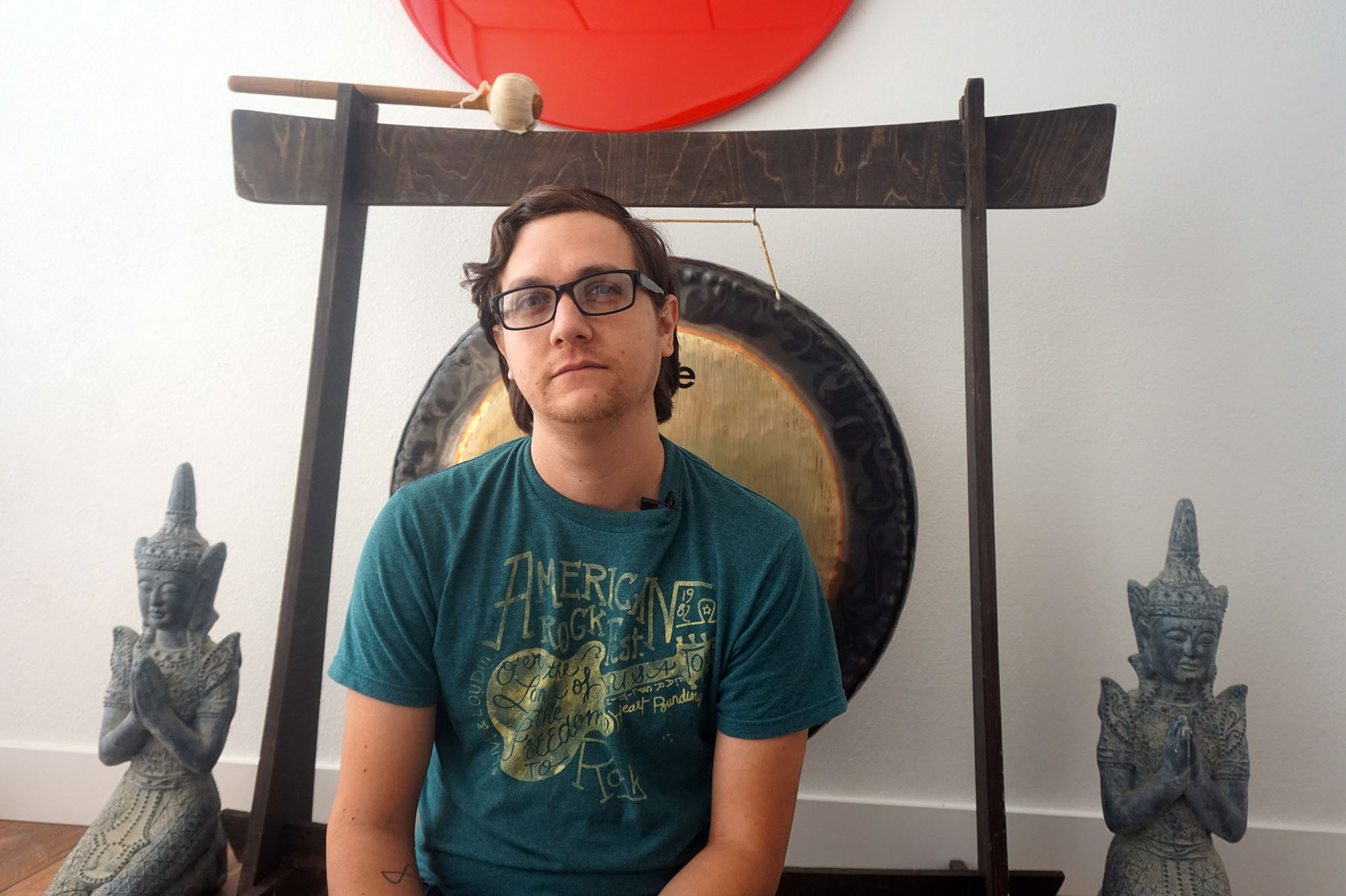 This is a photo of a man, Mitchell Maldonado. He is seated in front of a gong and a wall with a shiny red circle that is made to look like the flag of Japan. Mitchell is wearing glasses. He looks proud.