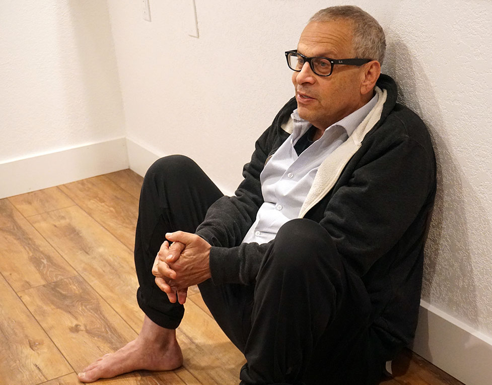 This is a photo of a man, Dr. Daniel Rieders, sitting on the floor. He has glasses and he is barefoot.