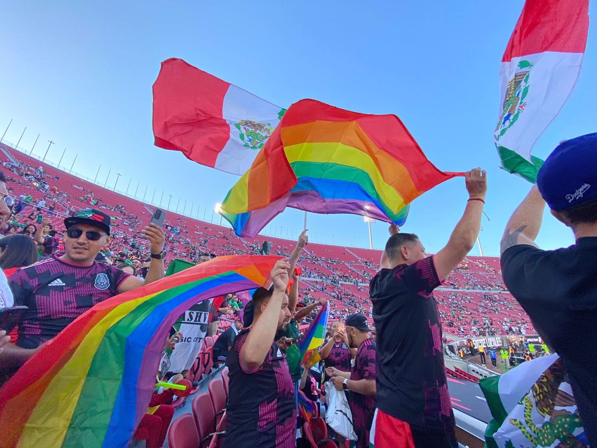 A crowd of soccer fans in a stadium wave Pride flags and Mexican flags.