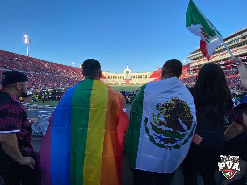 The backs of two audience members in a soccer stadium, one with a Pride flag draped over his shoulders and the other with a Mexican flag.