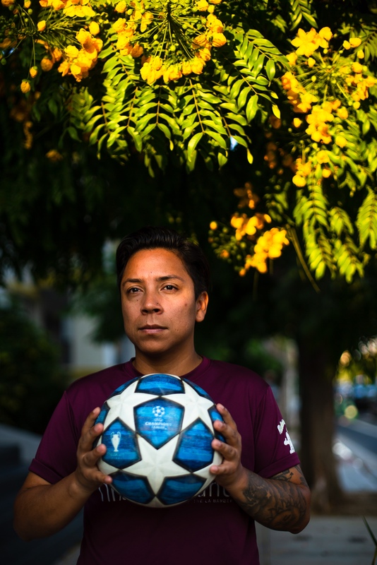 Gael stands on the sidewalk, holding a soccer ball in front of a tree.